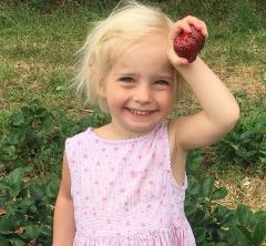 Artemis Alice Wood (girl, blonde) smiling and holding a strawberry