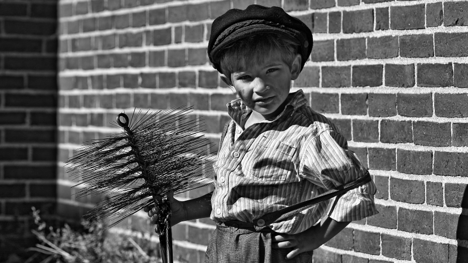 Black and white image of a young boy dressed in sooty clothes holding a chimney sweep