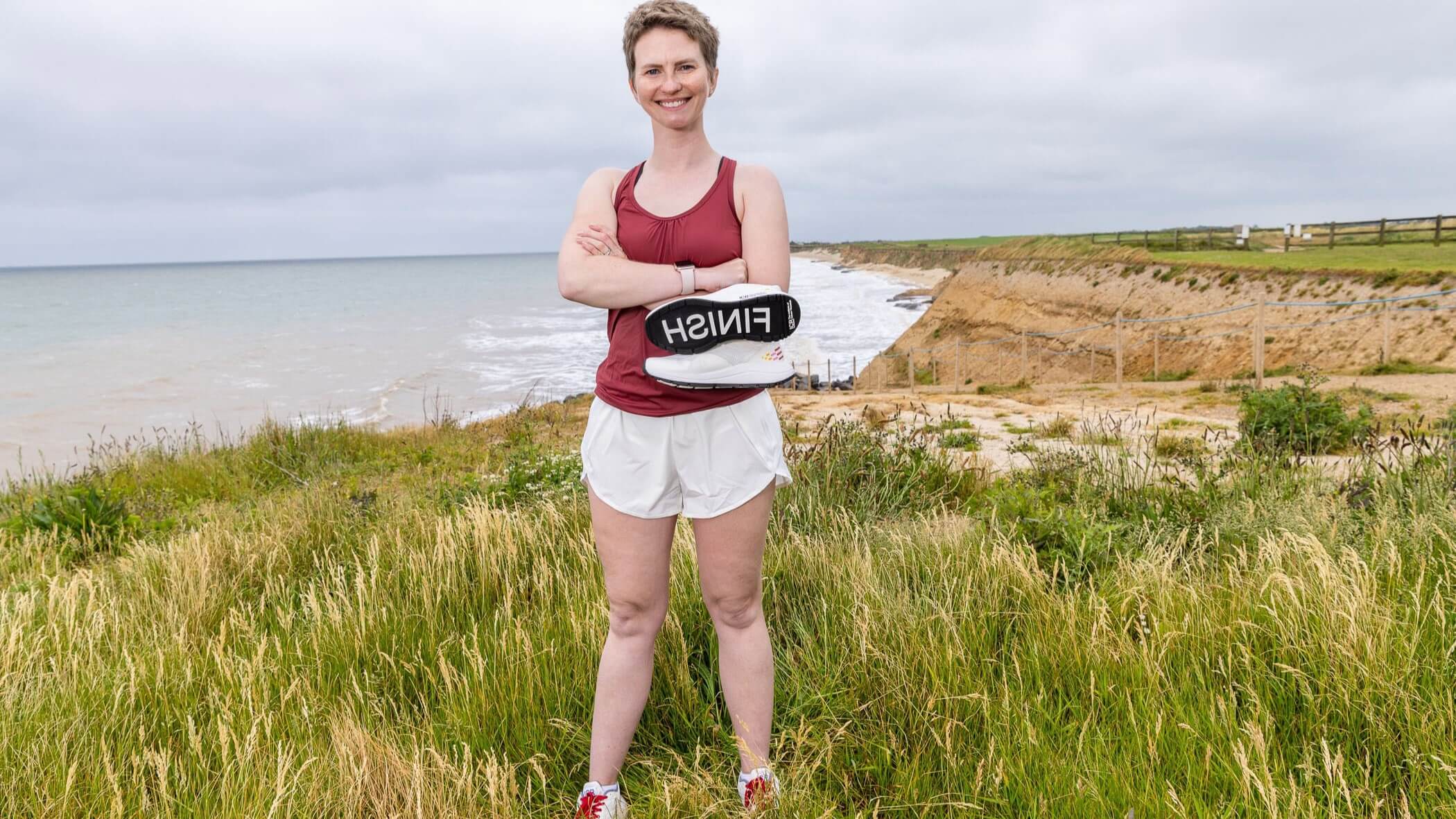Erin stands on a beach with her arms crossed holding a specially made ICR running shoe with FINISH CANCER on the sole