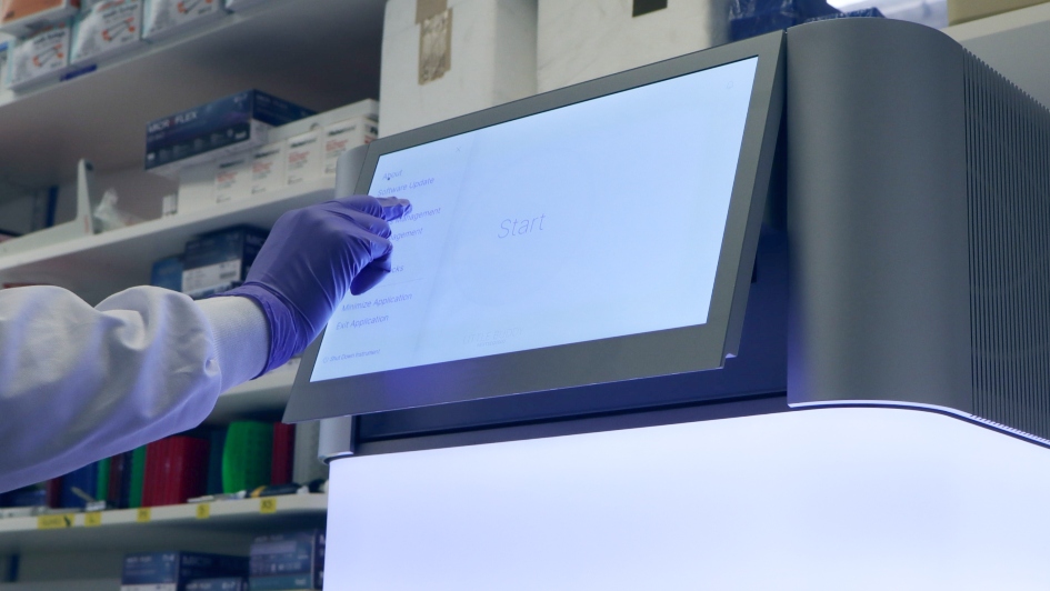 Close up of the NextSeq 2000, a bright, cube-shaped machine, and a hand stretched out to touch the machine's screen