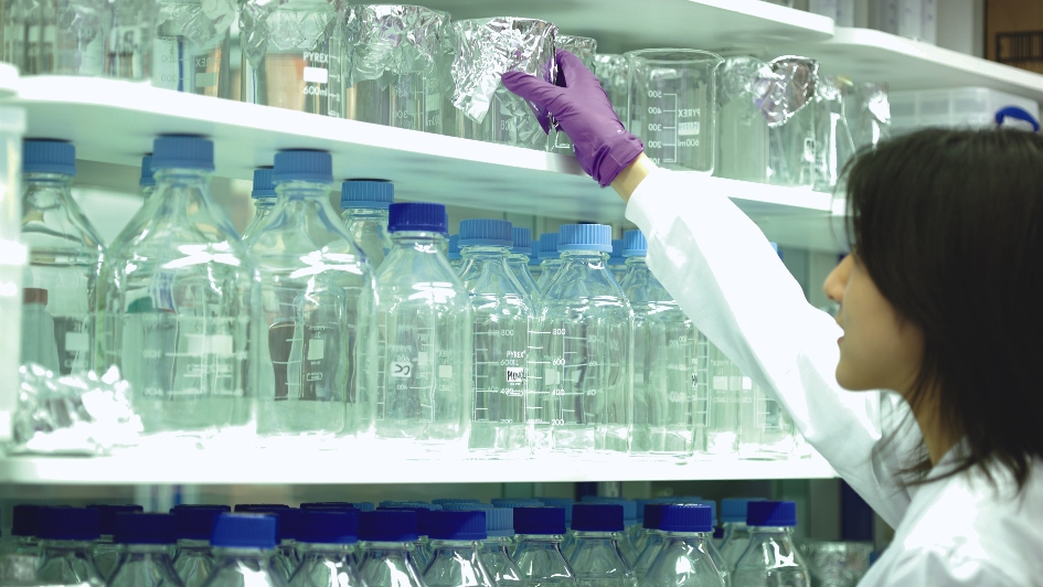 Person wearing a lab coat reaching for a glass beaker on a shelf filled with glass bottles and beakers