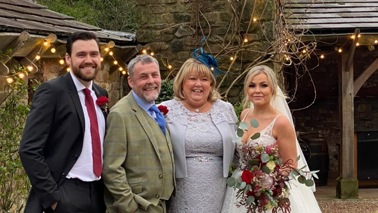 Glenys, her husband and son dressed smartly smile to the camera standing next to her daughter, a bride in a white dress and veil holding a floral bouquet