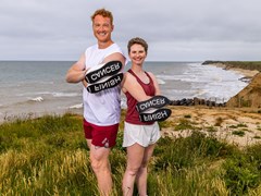 Sporting Champions Greg Rutherford (left) and Erin Kennedy (right) posing on a clifftop with the sea and beach behind them as they hold the custom trainers so you can see the words FINISH and CANCER on the soles.