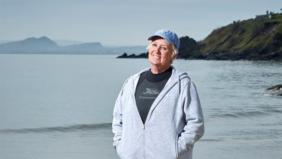 Karen stands on a beach with mountains in the background, wearing a grey hoodie and blue baseball cap