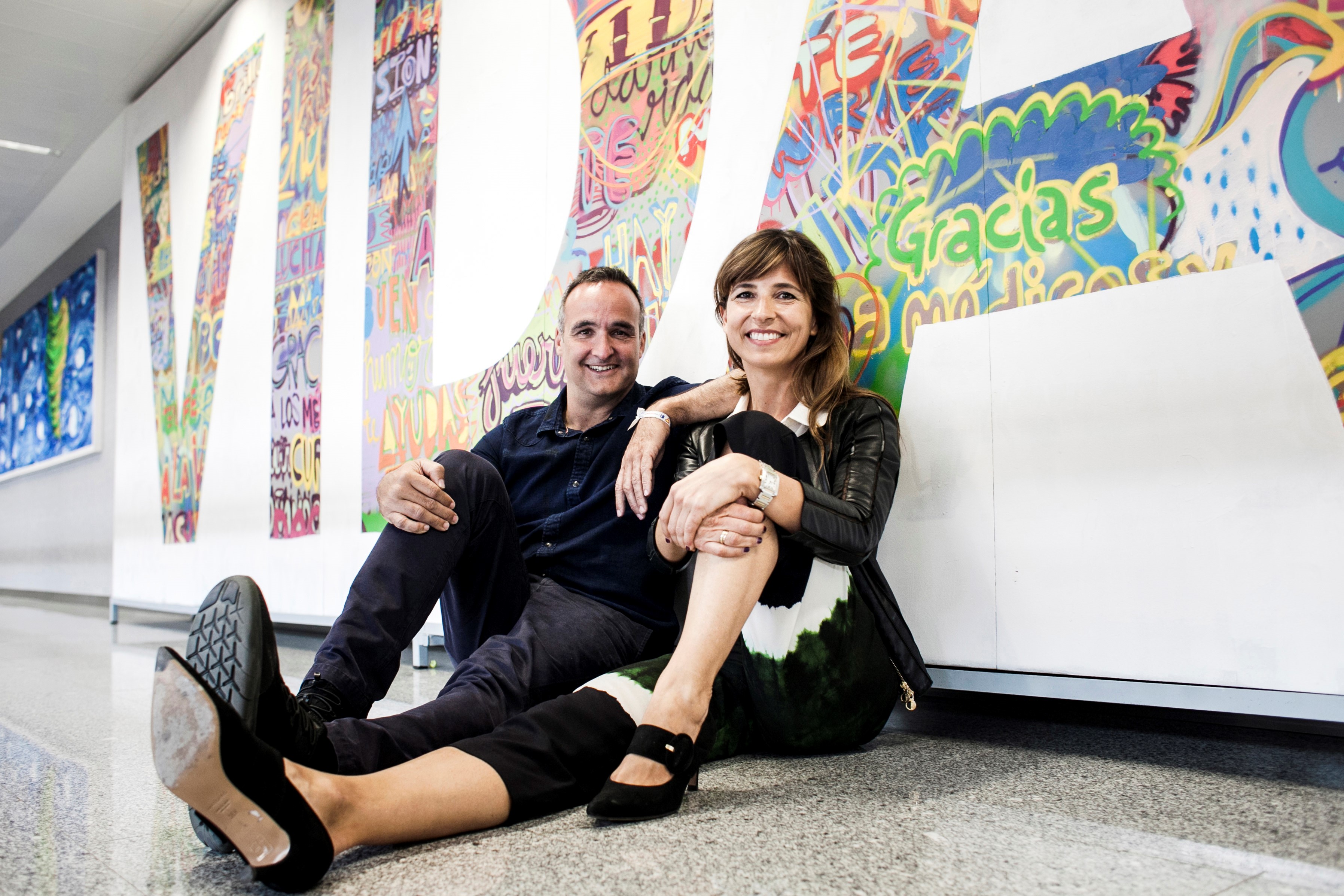 Lola and her husband Diego sit on an art gallery floor next to each other smiling at the camera