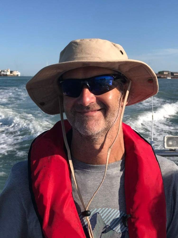 Mark wears a hat and sunglasses poses on a boat with the sea behind him