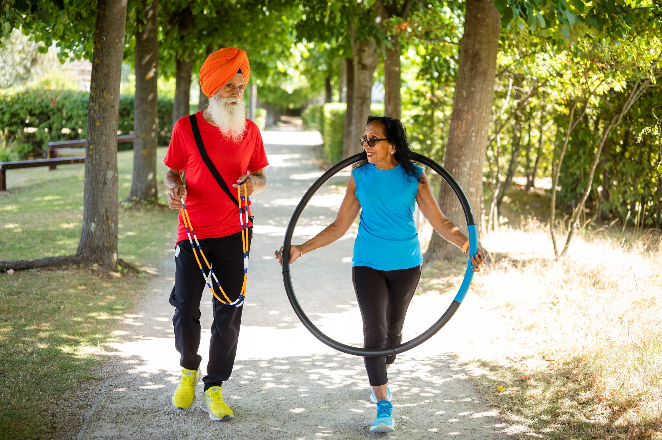 Pritpal and her husband walk along a tree-lined path. Pritpal wears a bright blue t shirt and sunglasses holds a hula hoop and smiles at her husband who wears an orange turban and carried a skipping rope